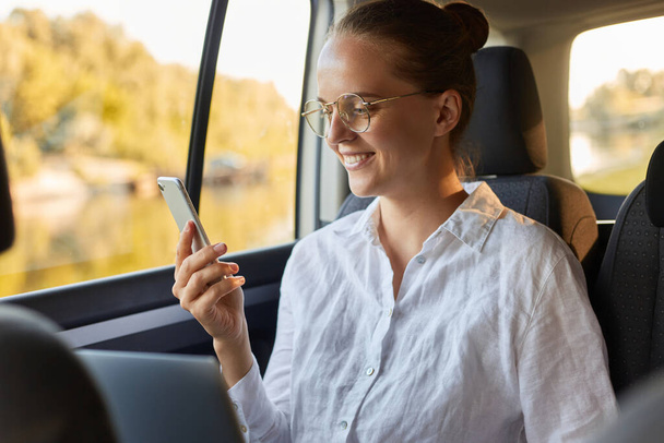 Portrait of happy optimistic woman in glasses and with bun hairstyle using laptop and mobile phone while sitting in the car, reading message on phone or checking social networks. - Zdjęcie, obraz