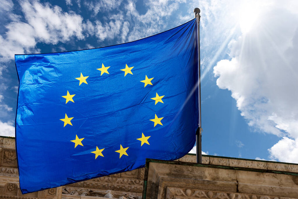 Primo piano di una bandiera dell'Unione europea che sventola contro un cielo azzurro con nuvole e raggi di sole. Brescia, Lombardia, Italia, Europa. - Foto, immagini