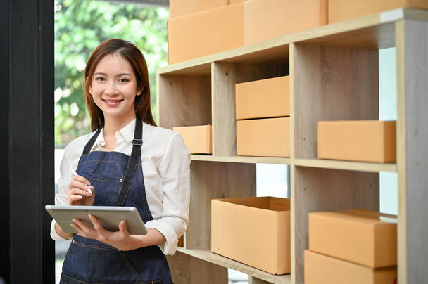 Beautiful attractive young Asian e-commerce business woman or online shop owner stands in her stock room with her portable digital tablet. - Photo, Image