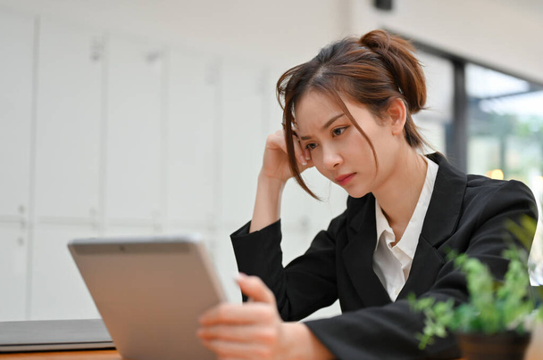 Przemyślana młoda azjatycka bizneswoman korzystająca z tabletu, kontemplująca strategię biznesową i koncentrująca się na swoim projekcie. - Zdjęcie, obraz