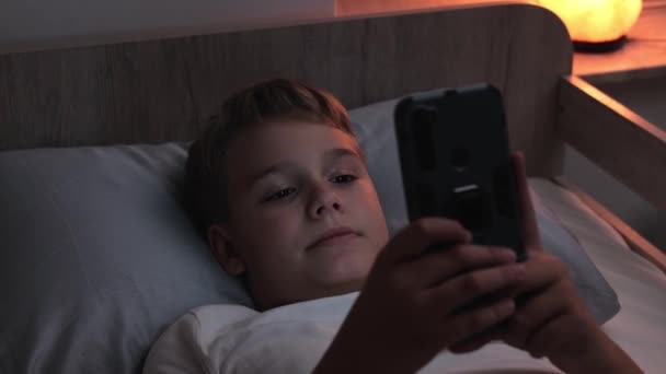 Teini-ikäinen käyttää älypuhelinta yöllä huonossa valaistuksessa nukkuessaan sängyssä. Käsite huonoja tapoja ja unettomuus - Materiaali, video