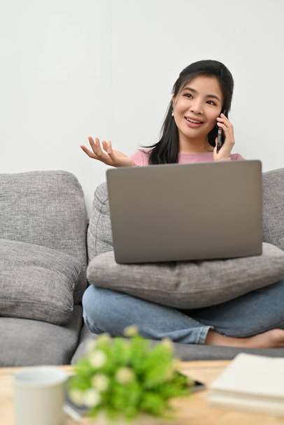 Portrait, attrayant millénaire asiatique femelle se détend dans le salon profiter de parler avec ses amis sur son téléphone tout en travaillant sur son ordinateur portable. - Photo, image