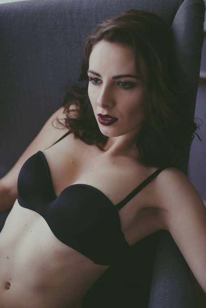 A young attractive girl with dark hair in black lingerie with evening make-up  sitting on a grey armchair  in the bedroom. beauty and fashion - Fotó, kép