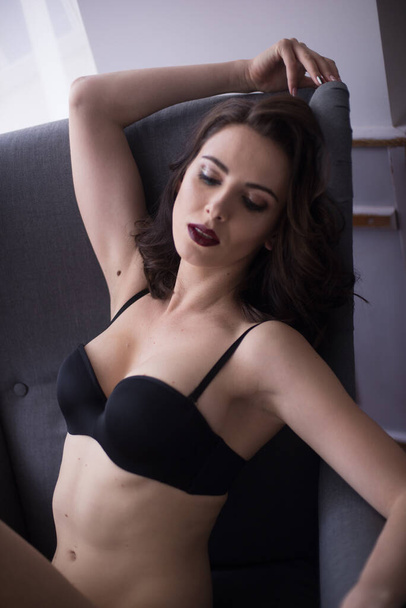 A young attractive girl with dark hair in black lingerie with evening make-up  sitting on a grey armchair  in the bedroom. beauty and fashion - Foto, imagen