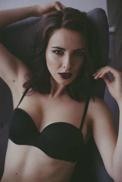 A young attractive girl with dark hair in black lingerie with evening make-up  sitting on a grey armchair  in the bedroom. beauty and fashion - Foto, Bild
