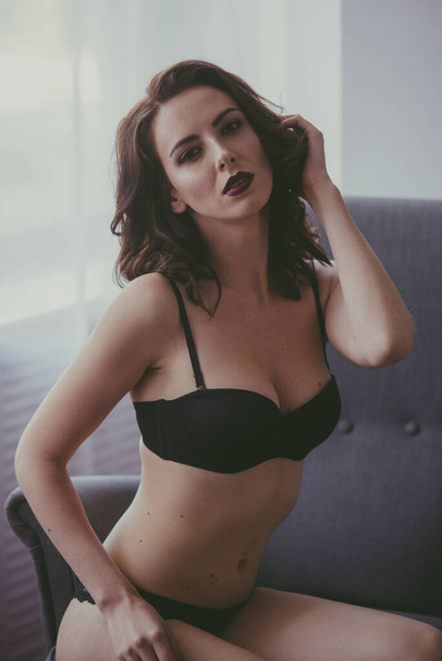 A young attractive girl with dark hair in black lingerie with evening make-up  sitting on a grey armchair  in the bedroom. beauty and fashion - Valokuva, kuva