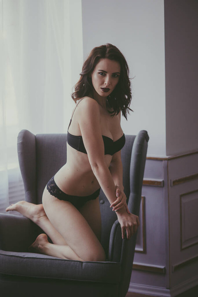 A young attractive girl with dark hair in black lingerie with evening make-up  sitting on a grey armchair  in the bedroom. beauty and fashion - Foto, immagini