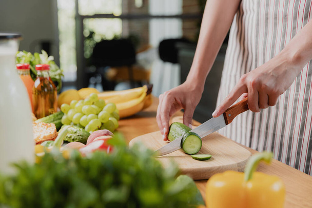 Close up on young housewife woman or girl hands slicing cucumber with kitchen knife, cuts food on wooden chopping board. Unrecognizable female standing at kitchen counter, cutting organic vegetables - Foto, Imagem