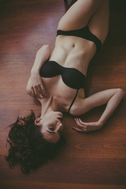 A young attractive girl with dark hair in black lingerie with evening make-up lie on the floor in the bedroom. beauty and fashion - Foto, Imagen