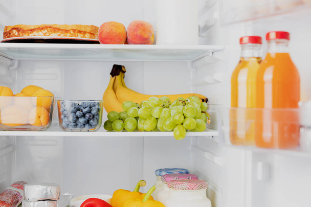 Vooraanzicht van open koelkast met twee deuren of koelkastdeur gevuld met vers fruit, groenten, sap, vol gezonde levensmiddelen en ingrediënten binnen. Elektrische keuken en huishoudelijke huishoudelijke apparaten - Foto, afbeelding