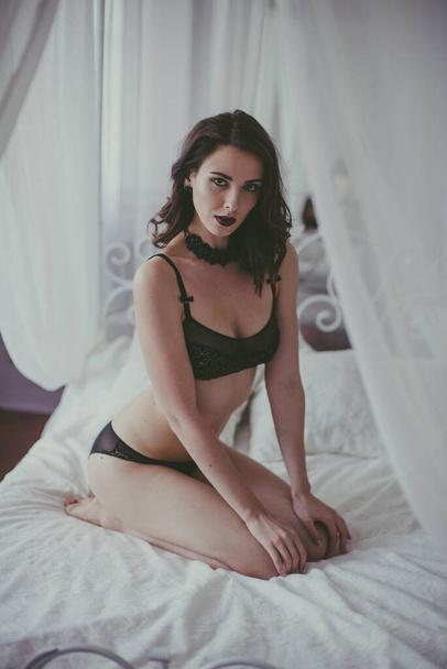 A young attractive girl with dark hair in black lingerie with evening make-up and jewelry and a white bathrobe on a white bed in the bedroom. beauty and fashion - Foto, imagen