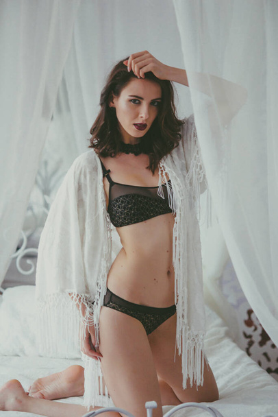 A young attractive girl with dark hair in black lingerie with evening make-up and jewelry and a white bathrobe on a white bed in the bedroom. beauty and fashion - Zdjęcie, obraz