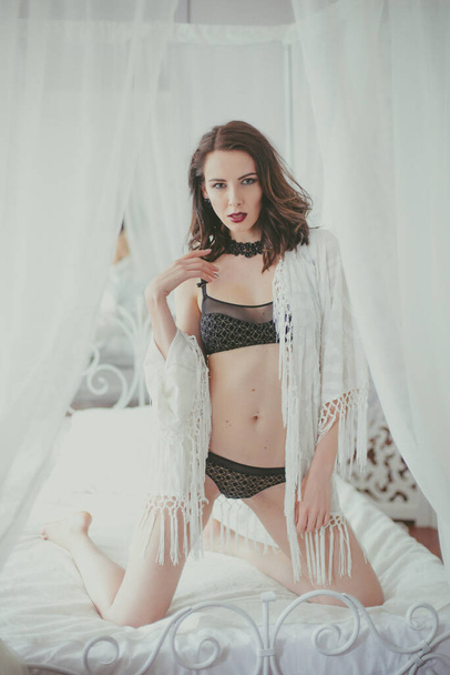 A young attractive girl with dark hair in black lingerie with evening make-up and jewelry and a white bathrobe on a white bed in the bedroom. beauty and fashion - Φωτογραφία, εικόνα