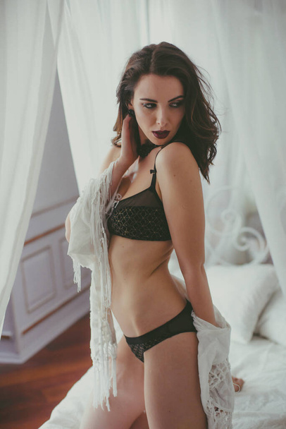 A young attractive girl with dark hair in black lingerie with evening make-up and jewelry and a white bathrobe on a white bed in the bedroom. beauty and fashion - Fotó, kép