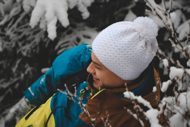 Πρωτότυπο πορτραίτο νεαρού χαμογελαστού άνδρα με πολύχρωμο μπουφάν χειμερινής πεζοπορίας, ζέσταμα και λευκό καπέλο κατά τη διάρκεια της χειμερινής περιόδου σε χιονισμένη περιοχή. Ένα αγόρι θαμμένο στο χιόνι. - Φωτογραφία, εικόνα