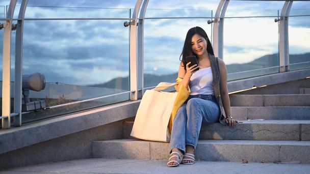 Pretty asian woman sitting using mobile phone and sitting on stairs at terrace with beautiful evening sky in background. - Photo, Image