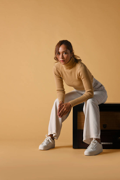 Attraktive modische Frau in trendigem Pullover sitzt auf einem Vintage-Radio, Studioaufnahme auf beigem Hintergrund. - Foto, Bild