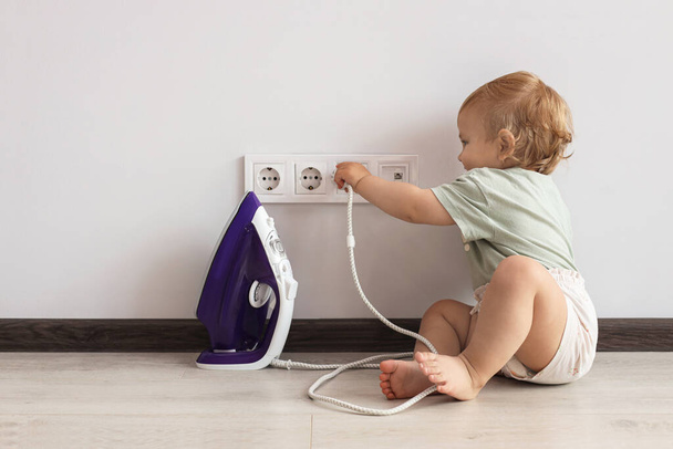 Cute baby playing with electrical socket and iron plug at home. Dangerous situation - Fotografie, Obrázek