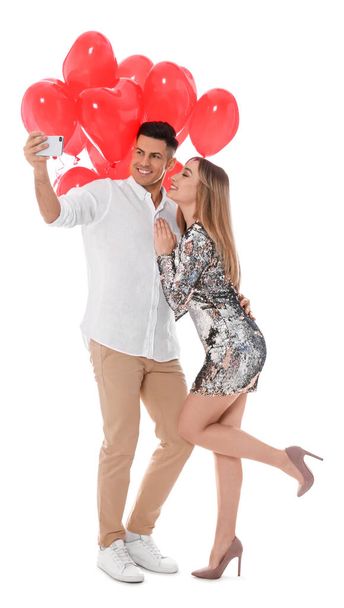 Lovely couple with heart shaped balloons taking selfie on white background. Valentine's day celebration - Фото, изображение