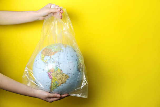 Woman holding globe in plastic bag against yellow background, closeup. Space for text. Environmental conservation - Fotoğraf, Görsel