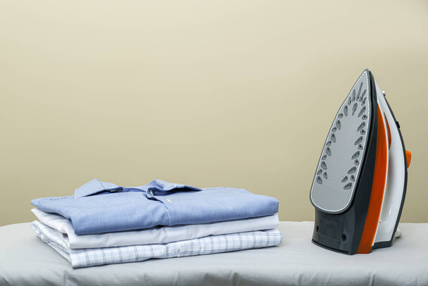 Modern iron and stack of clean clothes on board against beige background. Space for text - Фото, изображение