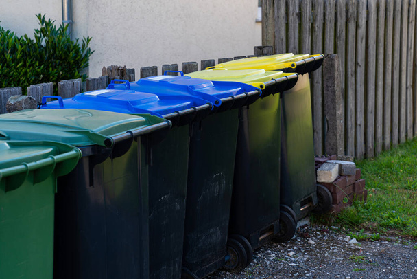 Ряд пластиковых мусорных баков вдоль забора. Многоцветная крышка. - Фото, изображение