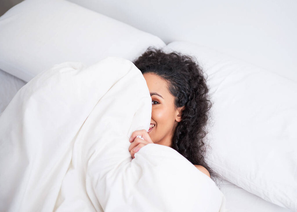 A young multi-ethnic woman hides playfully under the duvet in bed white linen. High quality photo - Foto, Imagen