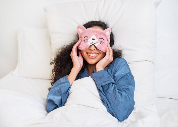 Uma jovem mulher multi-étnica acorda na cama com uma máscara de sono bonito gatinho. Foto de alta qualidade - Foto, Imagem