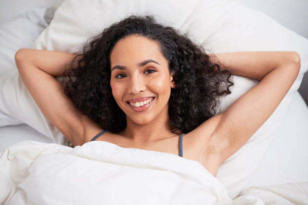 Egy fiatal, többnemzetiségű nő fekszik karokkal a tarkóján az ágyban, mosolyogva. Kiváló minőségű fénykép - Fotó, kép