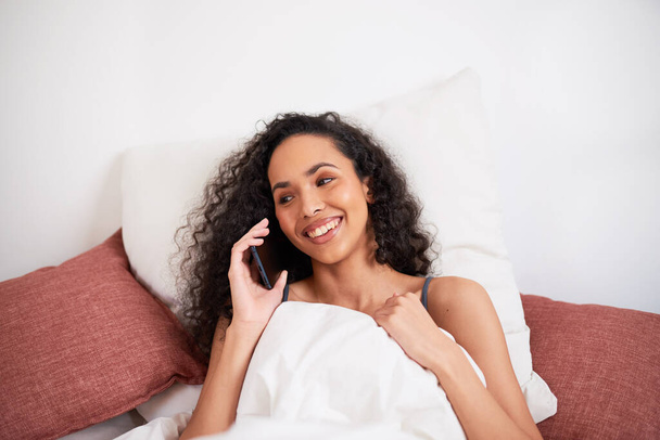 Gros plan d'une jeune femme multi-ethnique bavardant sur son téléphone portable au lit avec des oreillers. Photo de haute qualité - Photo, image