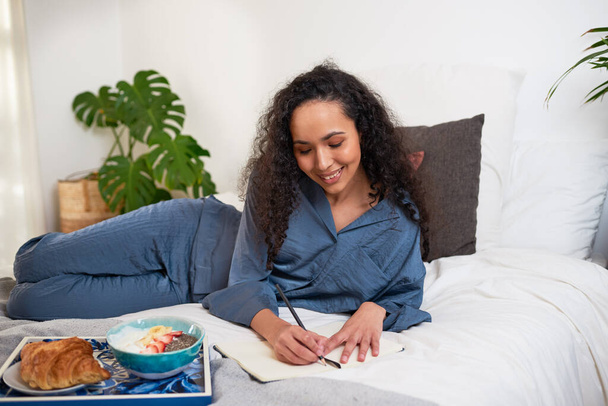 Egy fiatal nőnek van egy önellátó napja az ágyban, reggelizik és naplót ír. Kiváló minőségű fénykép - Fotó, kép