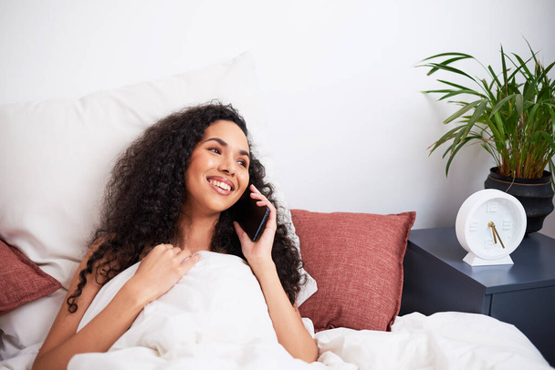 Молодая многонациональная женщина смеется во время интимного междугороднего звонка с кровати. Высокое качество фото - Фото, изображение