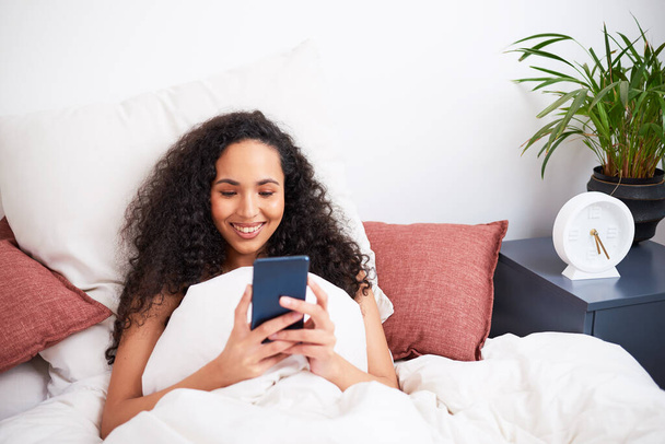 Een jonge multi-etnische vrouw zit in bed met kussens op haar mobieltje. Hoge kwaliteit foto - Foto, afbeelding
