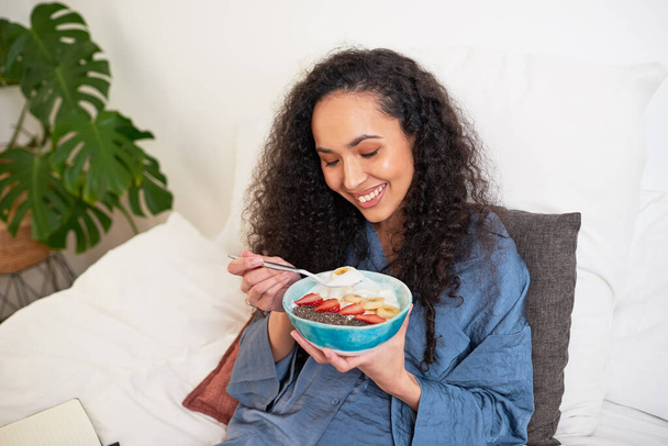 A beautiful multi-ethnic woman eats fruit bowl in bed. High quality photo - Φωτογραφία, εικόνα