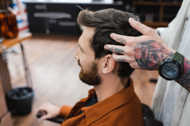 tattooed hairdresser in wristwatch touching hair of brunette man in barbershop - Valokuva, kuva
