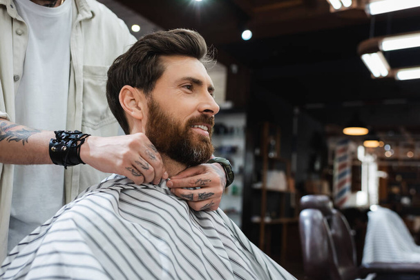 barber putting hairdressing cape on smiling bearded man  - Фото, изображение