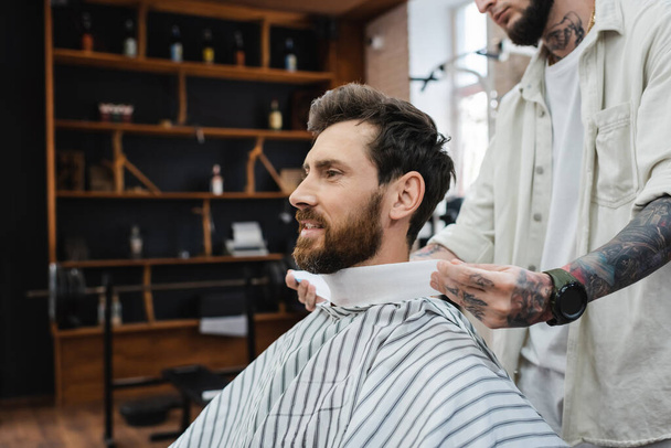 татуированный парикмахер обертывая шею бородатого мужчину с парикмахерским воротником - Фото, изображение