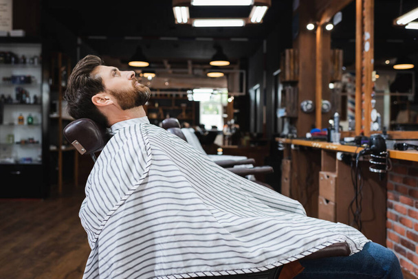 side view of brunette bearded man sitting in hairdressing cape in beauty salon - Foto, Imagen