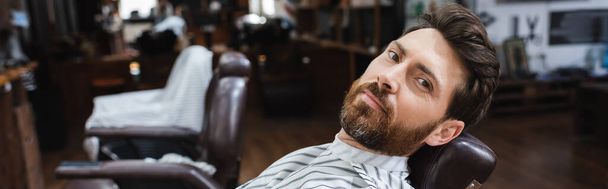 hombre barbudo mirando a la cámara mientras está sentado en la peluquería borrosa, pancarta - Foto, imagen