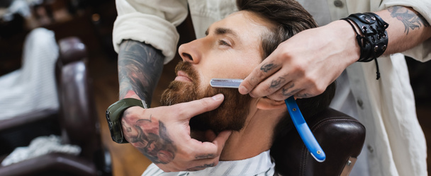 tattooed hairstylist in leather bracelet shaving man with straight razor, banner - Photo, Image