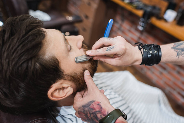 high angle view of tattooed barber shaving bearded man with straight razor - Fotoğraf, Görsel