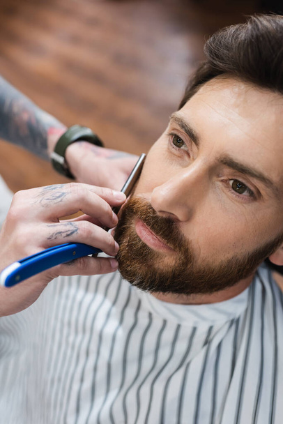 Hochwinkelaufnahme eines bärtigen Mannes in der Nähe des Friseurs, der ihn mit einem Rasiermesser rasiert - Foto, Bild