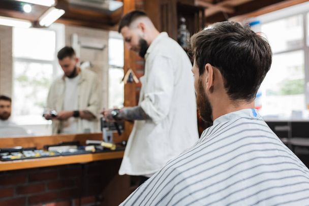 brunette man in hairdressing cape near blurred mirror and hairstylist in barbershop - Foto, Bild