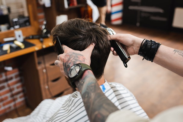 barbier tatoué dans la montre-bracelet tenant tondeuse à cheveux et peigne près de l'homme brune - Photo, image