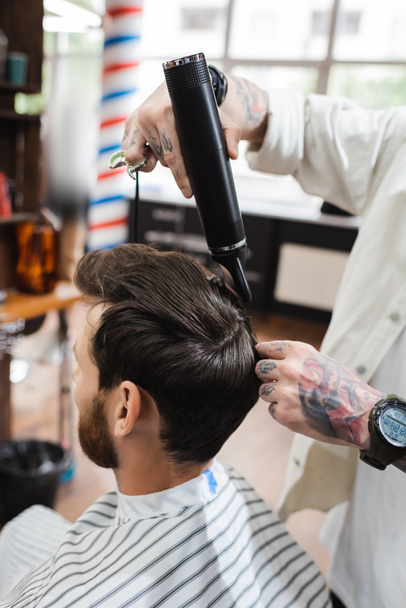 Kol saatinde dövmeli kuaför berber dükkanında müşterinin saçını kurutuyor. - Fotoğraf, Görsel