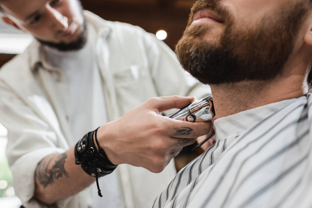partial view of blurred barber shaving neck of bearded man with electric razor - Photo, Image