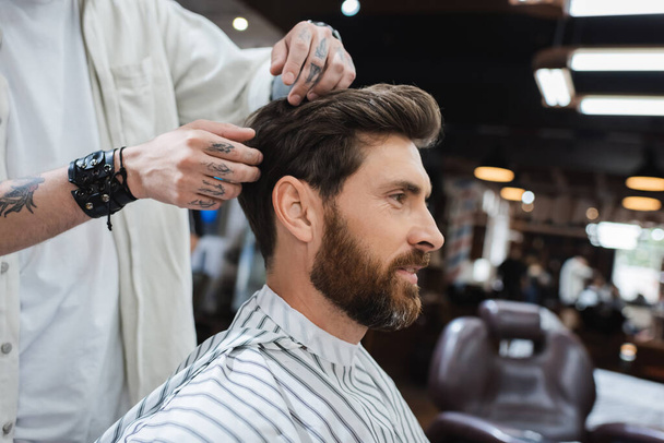 morena barbudo hombre en peluquería capa cerca de peluquero tatuado tocando su pelo en salón de belleza - Foto, Imagen