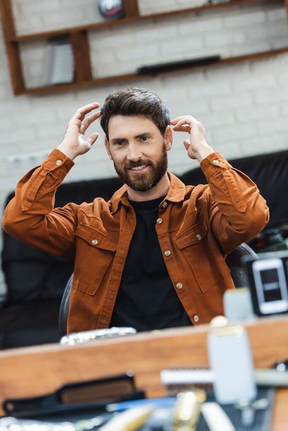 pleased bearded man looking in mirror and touching hair in beauty salon - Photo, Image
