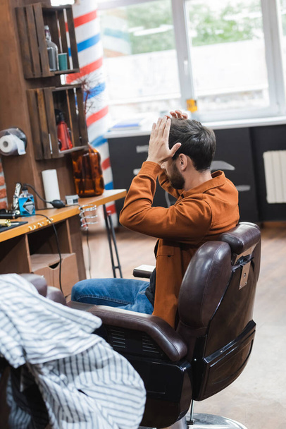 brunette homme toucher les cheveux tout en étant assis dans la chaise de coiffeur dans le salon de beauté - Photo, image