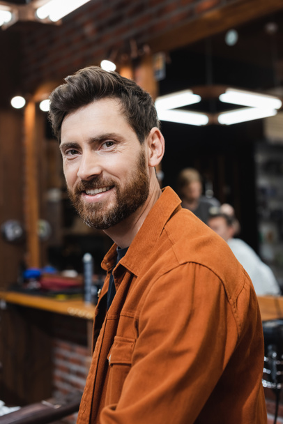 brunette homme avec barbe souriant à la caméra dans barbier flou - Photo, image